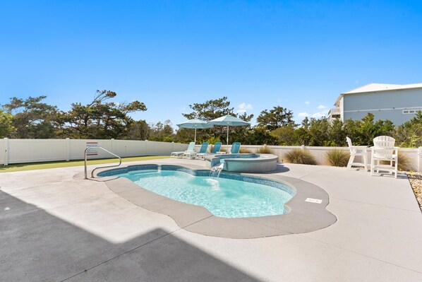 A swimming pool with lounge chairs and umbrellas, surrounded by a white fence and trees.