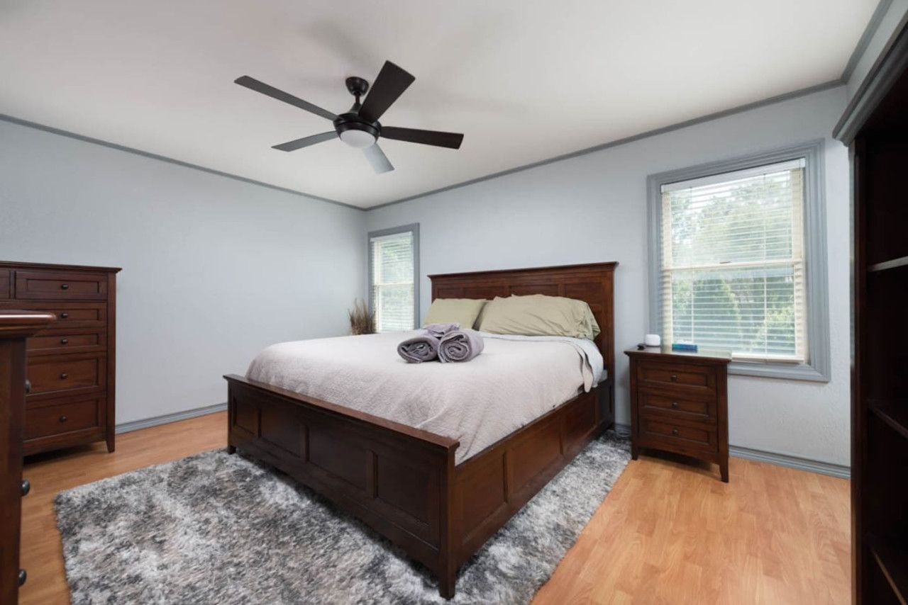 Een slaapkamer met een houten bed, een plafondventilator, een dressoir, een nachtkastje en een raam met jaloezieën.