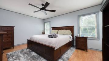 Een slaapkamer met een houten bed, een plafondventilator, een dressoir, een nachtkastje en een raam met jaloezieën.