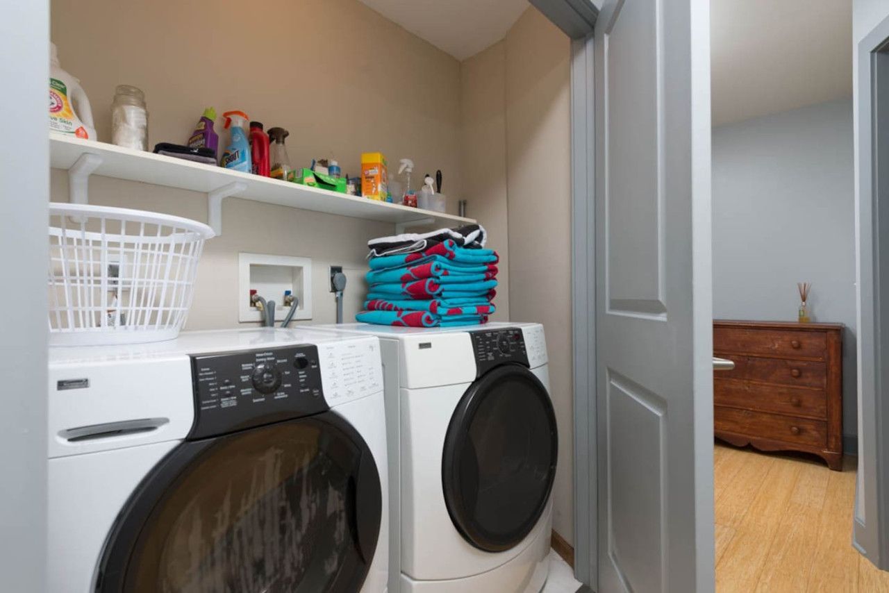 Een wasruimte met twee wasmachines, een mand en planken met schoonmaakmiddelen.