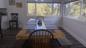 Un espace repas avec une table et des bancs en bois, de grandes fenêtres donnant sur la forêt, et un coin cuisine équipé d’un évier et de placards.