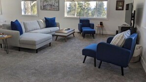 Un salon moderne avec un canapé d’angle gris, des fauteuils bleus et une table basse. De grandes fenêtres laissent entrer la lumière naturelle et offrent une vue sur les arbres.