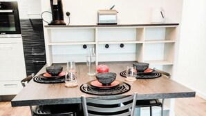 Et moderne spiseområde med et rektangulært bord dækket til fire personer, omgivet af metalstole. En kaffemaskine og et lille apparat er synlige på væggen over bordet.