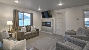 Een woonkamer met een grijze bank, gestreept fauteuils, een flatscreen televisie, een open haard en een raam met gordijnen.