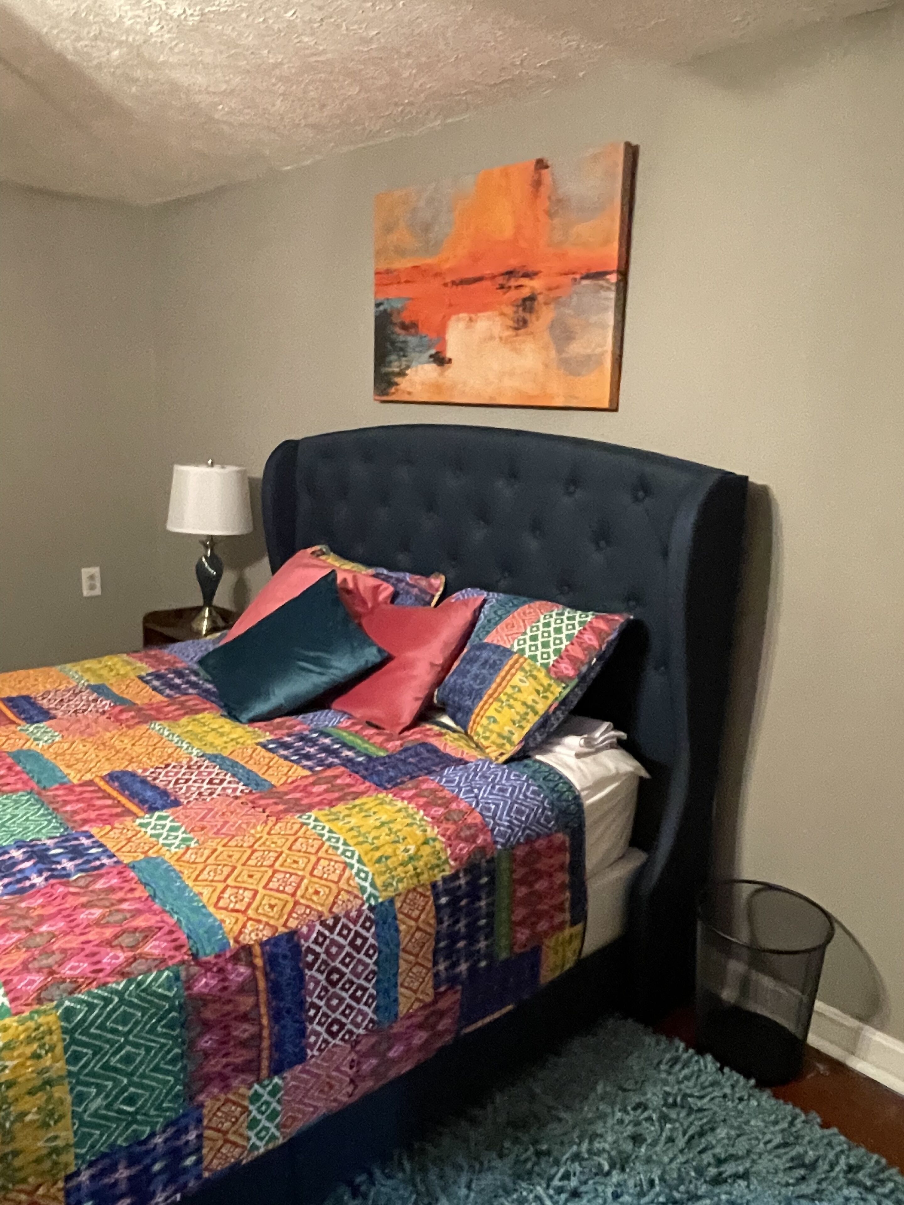 Une chambre à coucher avec une tête de lit capitonnée, une courtepointe aux couleurs vives, un tapis bleu, une lampe de chevet et un tableau accroché au mur.