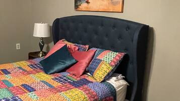 Une chambre à coucher avec une tête de lit capitonnée, une courtepointe aux couleurs vives, un tapis bleu, une lampe de chevet et un tableau accroché au mur.