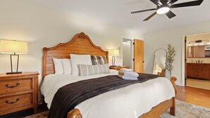 Un dormitorio con cama de madera, lámparas de noche, ventilador de techo y baño visible a través de una puerta abierta.
