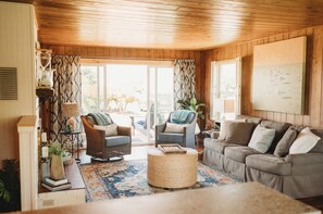 A cozy living room with wooden walls and ceiling, a large window with patterned curtains, a grey sofa, two wicker chairs, and a coffee table.