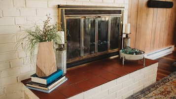 Una chimenea con repisa de madera, una planta en maceta y una pila de libros sobre un muro de ladrillo blanco.