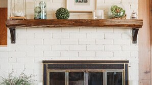 Una chimenea con pared de ladrillo blanco, repisa de madera y varios objetos decorativos, incluyendo una obra de arte enmarcada, una piña y recipientes de vidrio.
