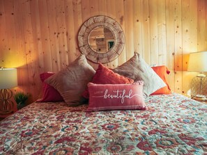 Ein Schlafzimmer mit einer Holzwand, einem runden Spiegel, floralem Bettbezug und einem Kissen mit dem Schriftzug 'LIFE IS beautiful'.