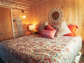 A cozy bedroom with a floral bedspread, a round woven mirror, and a wall-mounted macrame piece.