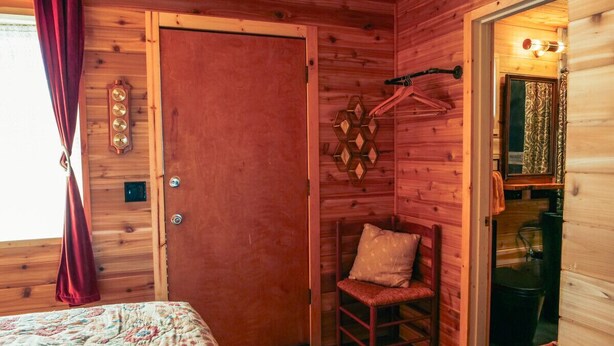 Ein Zimmer in einer Holzhütte mit einem Bett, einem Sessel, einer Tür und einem kleinen Badezimmer, das durch eine geöffnete Tür sichtbar ist.
