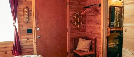 Habitación de cabaña de madera con cama, una silla, una puerta y un baño pequeño visible a través de una puerta abierta.