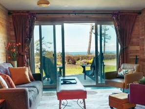 A modern living room with a sofa, chairs, and a coffee table. It features large glass doors leading to a deck with a view of the ocean. - #2 travel room · 2-Hot tub-Water view-Full kitchen-beach-1 bedroom (Sequim)