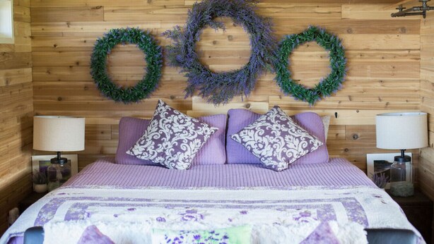 A bedroom with a wooden wall, two nightstands with lamps, a bed with purple bedding, and three wreaths on the wall. - Rm 4 Sequim lavender · 4-Water View-Beach access-Sequim's Lavender Room (Sequim)