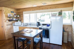 Une cuisine compacte aux murs en bois, avec un réfrigérateur blanc, une cuisinière et un évier. On y trouve des tabourets de bar et une petite table sur laquelle est posé un plateau contenant divers objets.