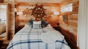 Une chambre à coucher aménagée dans une cabane en bois, avec un lit, des oreillers, un gouvernail de bateau fixé au mur et une fenêtre.