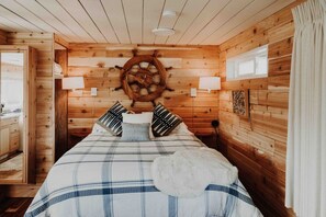 Une chambre à coucher aménagée dans une cabane en bois, avec un lit, des oreillers, un gouvernail de bateau fixé au mur et une fenêtre.