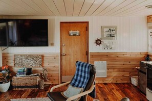 A cozy living room with a wooden floor, a television mounted on the wall, a door with a small shelf, and a kitchen area with a stove. - 5- Hot Tub & Massage Chair- Water view Cottage (sequim)