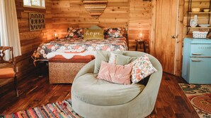 A cozy bedroom with a wooden interior, a bed with floral bedding, a vintage blue dresser, and a comfortable armchair with cushions.