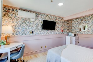 Un dormitorio con papel tapiz floral, una cama, un escritorio con silla, un televisor, un espejo y un aparador.
