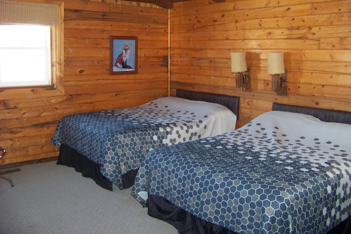 Ein Zimmer in einer Holzhütte mit zwei Betten, einem Fenster und einem gerahmten Bild an der Wand.