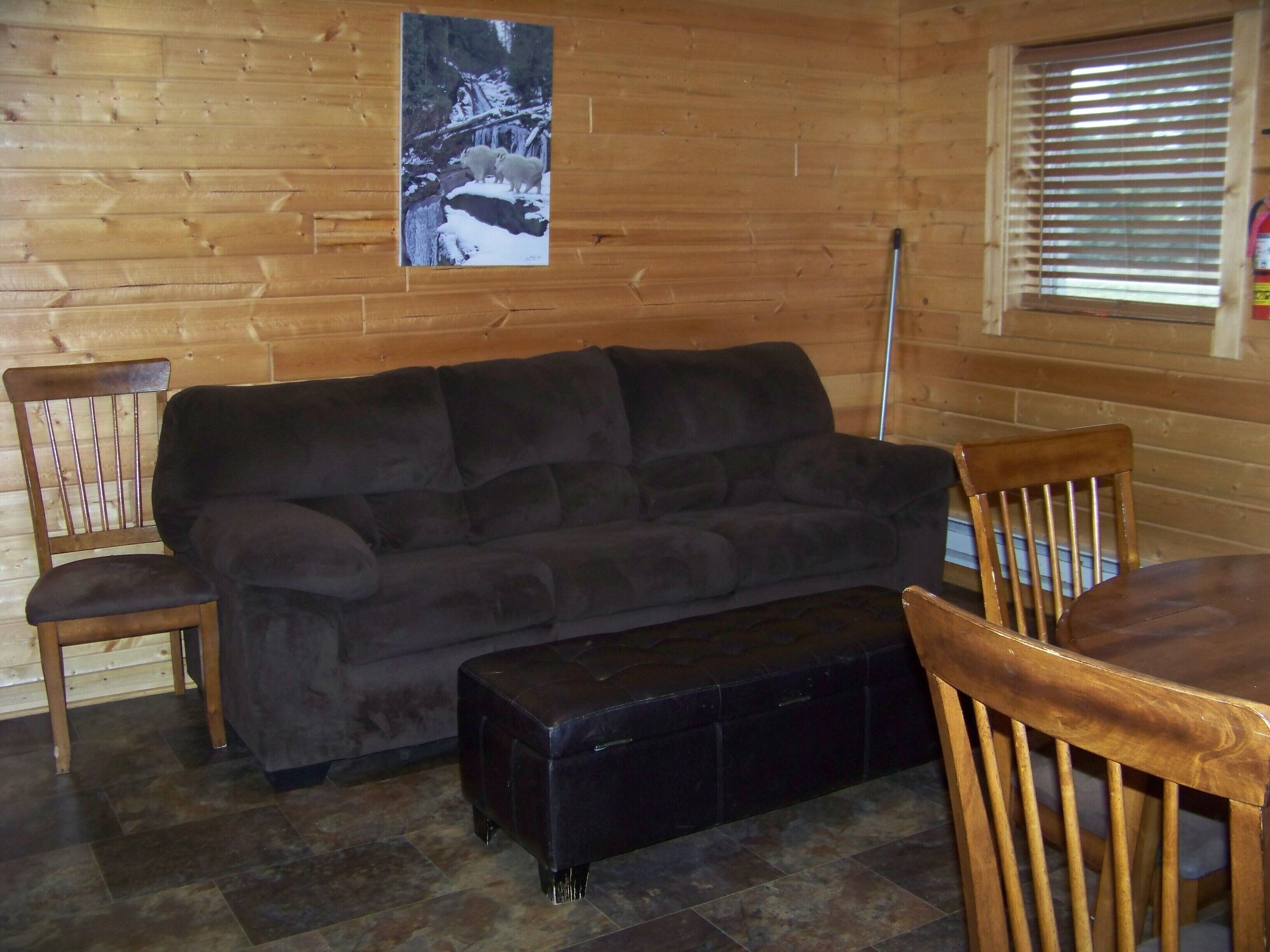 A cozy room with a dark grey sofa, wooden chairs, a small black ottoman, and a wooden floor.