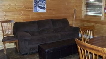 A cozy room with a dark grey sofa, wooden chairs, a small black ottoman, and a wooden floor.
