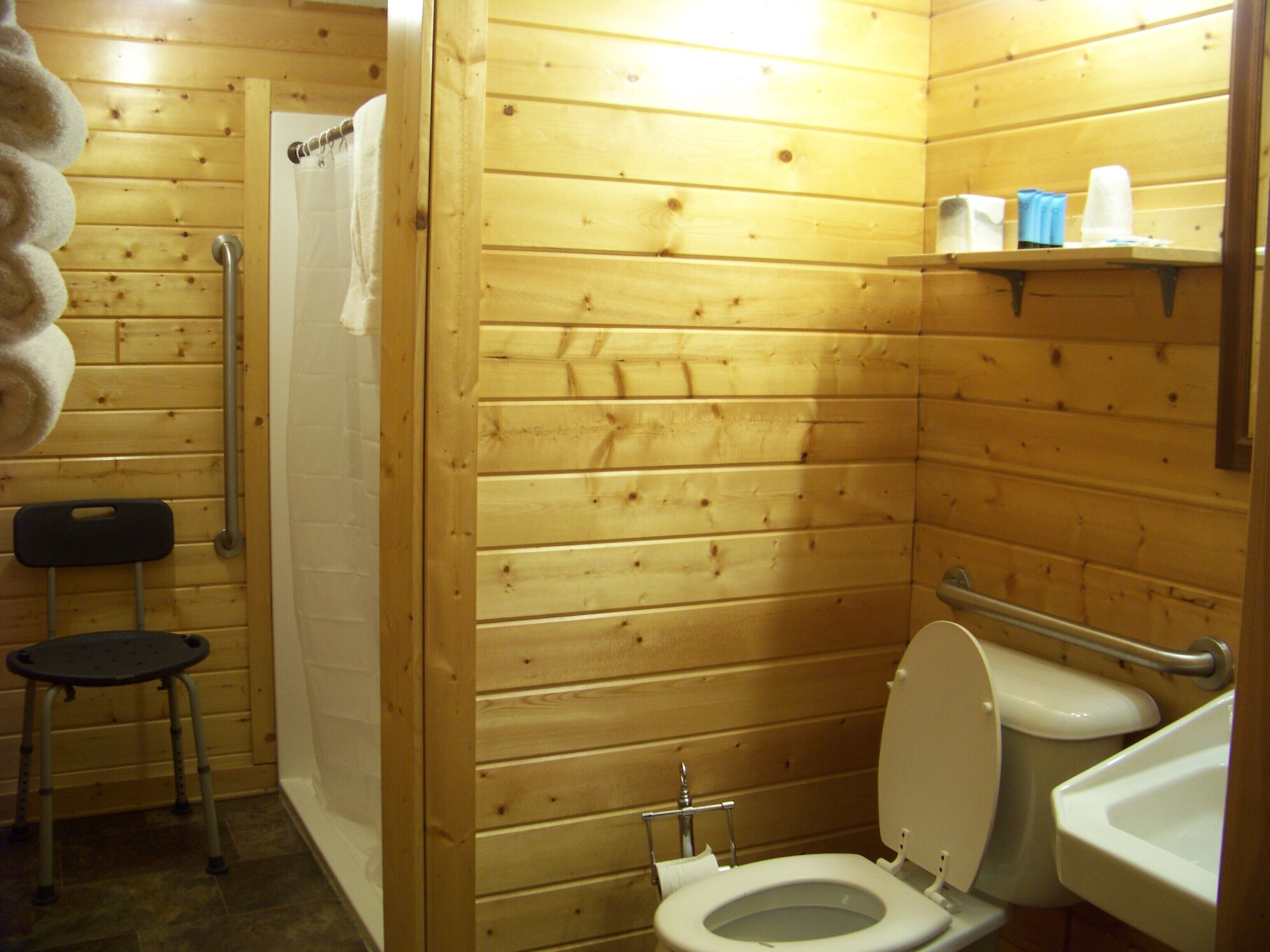 Une salle de bain aux murs lambrissés, équipée de toilettes, d’une douche et d’une petite chaise.