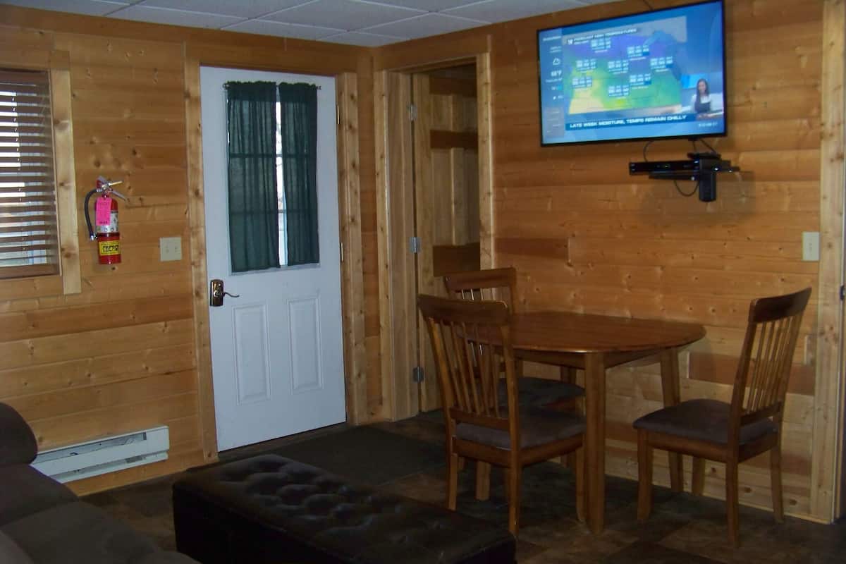 Ein gemütliches Zimmer mit Holzvertäfelung, einem an der Wand befestigten Fernseher, einer Tür mit Fenster und einer Essecke mit Stühlen und Tisch.