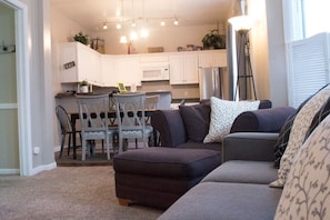 A living room with a dark-colored sofa, a dining table with chairs, and a kitchen area with white cabinets and a microwave. - Heart of Downtown 2-Bedroom Apt, Mountain Views, Full Kitchen, Walk to Eats (Glenwood Springs)