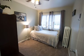 A bedroom with a bed, a radiator, a window with curtains, a ceiling fan, and a wall-mounted picture. - Heart of Downtown 2-Bedroom Apt, Mountain Views, Full Kitchen, Walk to Eats (Glenwood Springs)