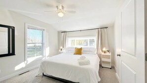 Ein geräumiges Schlafzimmer mit einem großen Bett, einem Deckenventilator, einem Fenster mit Vorhängen und einem Nachttisch mit Lampe.