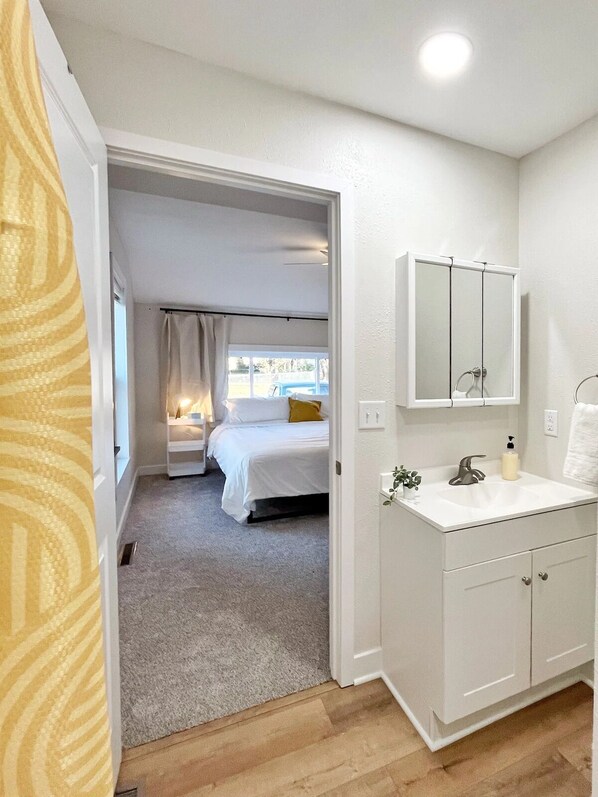 A bedroom with a bed, a bathroom with a sink and mirror, and a hallway leading to a balcony. - Katy Trail Rest Stop (Sedalia)