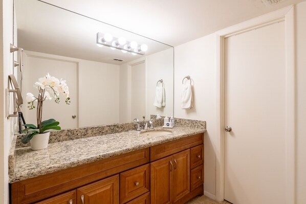 A bathroom with a granite countertop, wooden cabinets, a sink, a mirror with lights, and a potted orchid.