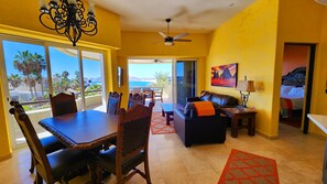A bright living room with a dining area, a sofa, a coffee table, and a TV. There is a balcony with a view of palm trees and the ocean.