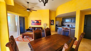 A dining area with a wooden table and chairs, a sofa, a ceiling fan, and a kitchen in the background.