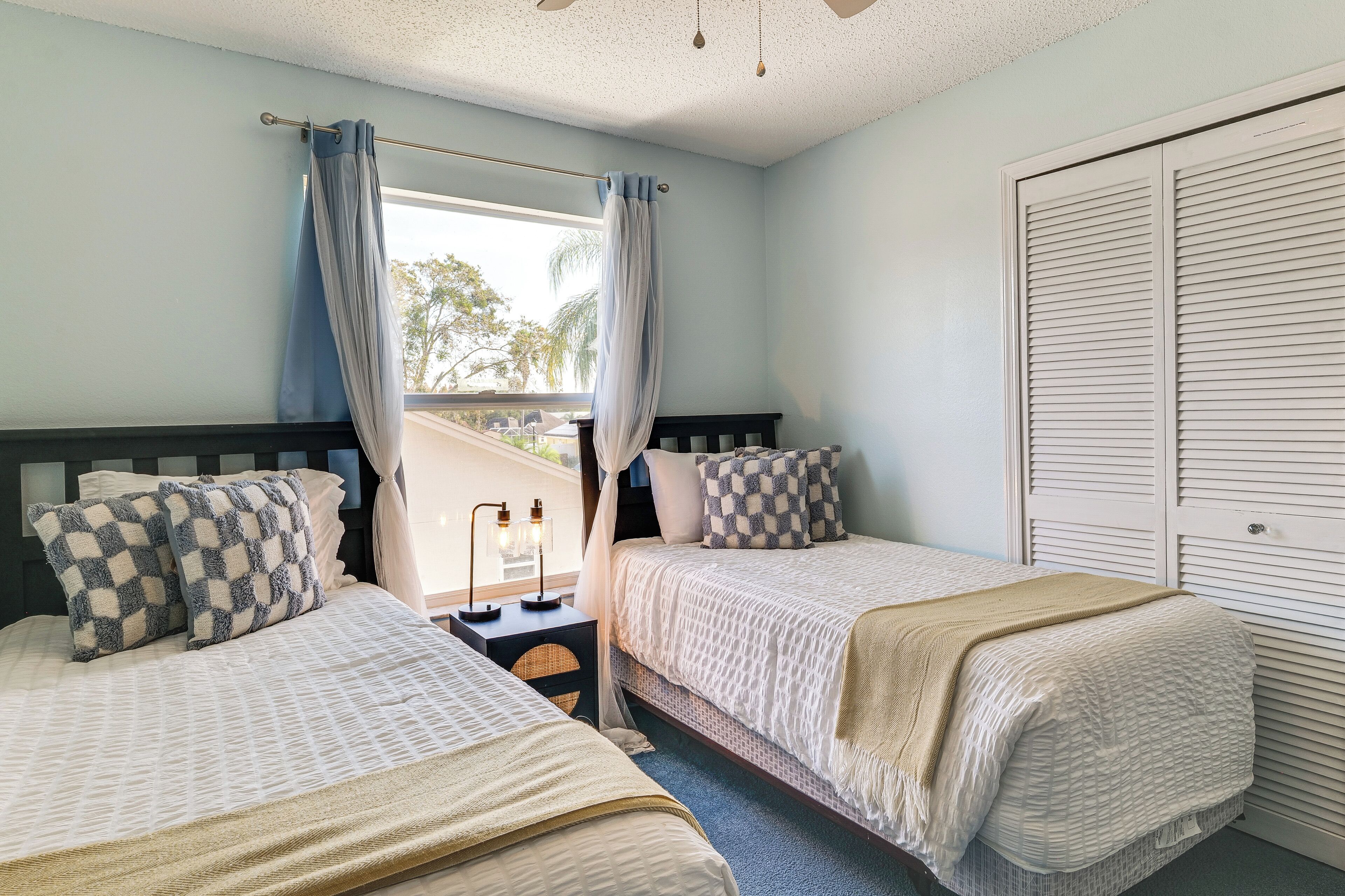 A bedroom with two beds, a nightstand with a lamp, a window with curtains, and a closet with louvered doors.
