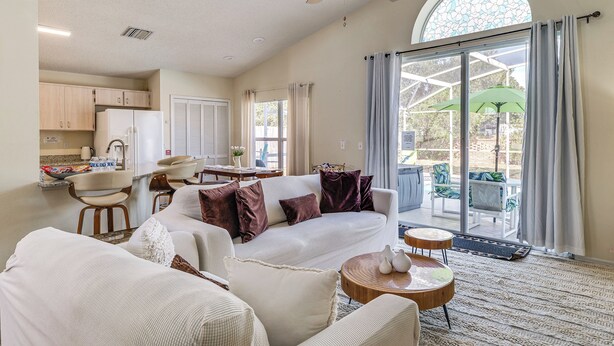 A spacious living room with a large white sofa, a dining area with a table and chairs, a kitchen with cabinets and appliances, and a sliding glass door leading to an outdoor patio with chairs and an umbrella. - 5 Mi to Disney! Family Getaway w/ Poolside Oasis (Kissimmee)