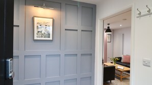 En gang med en grå dør med panel, et billede i ramme på væggen, og et glimt af et spiseområde med bord og stole.