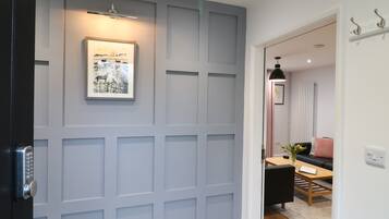 Un couloir avec une porte à panneaux gris, un tableau encadré au mur, et un aperçu d’un coin repas avec une table et des chaises.