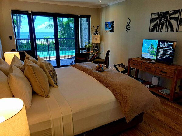 A bedroom with a large bed, a desk with a computer, a TV, and a view of the ocean through the sliding glass doors.