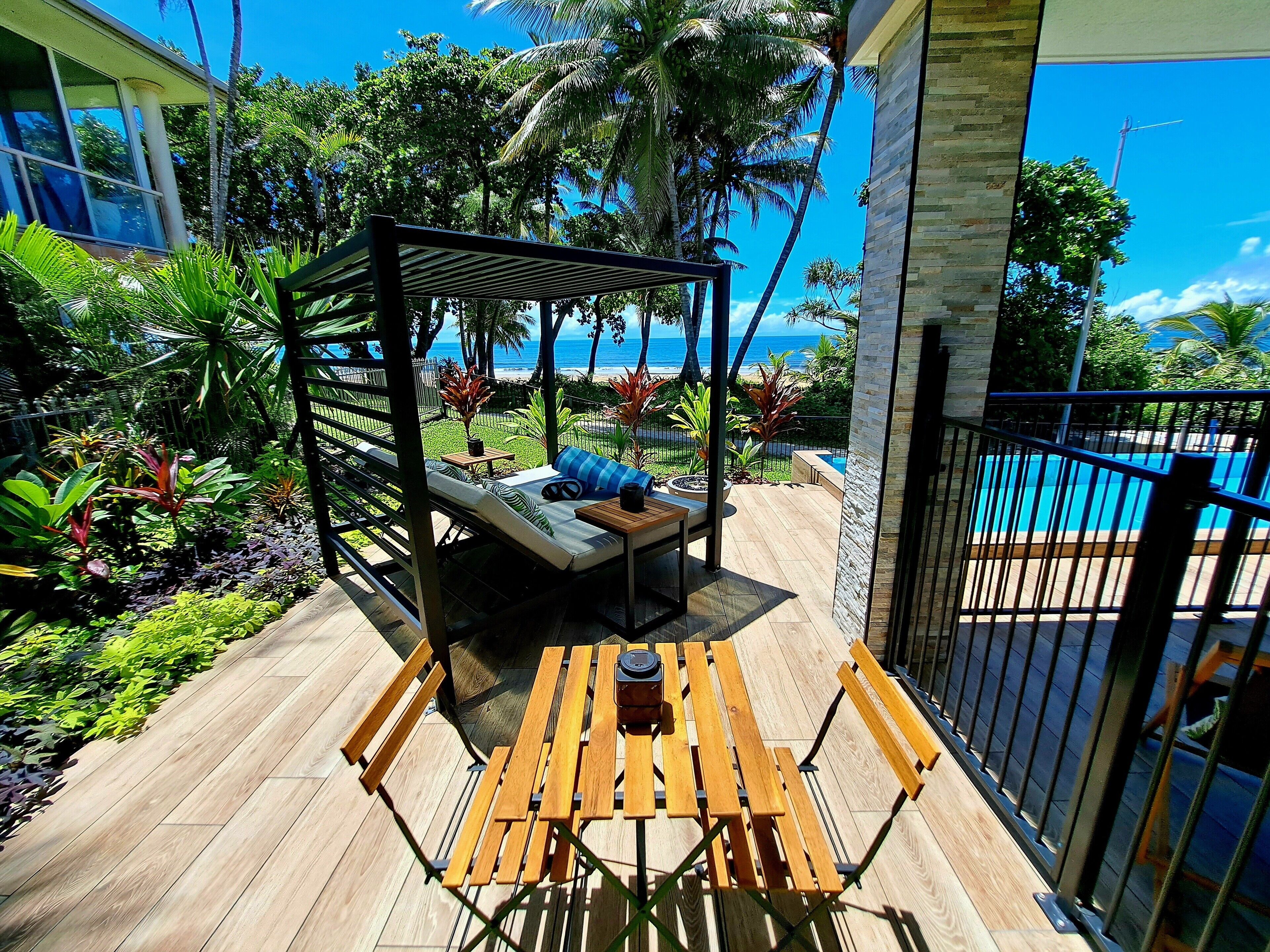 A wooden deck with a wooden bench and a daybed with pillows, surrounded by tropical plants and a view of the ocean.