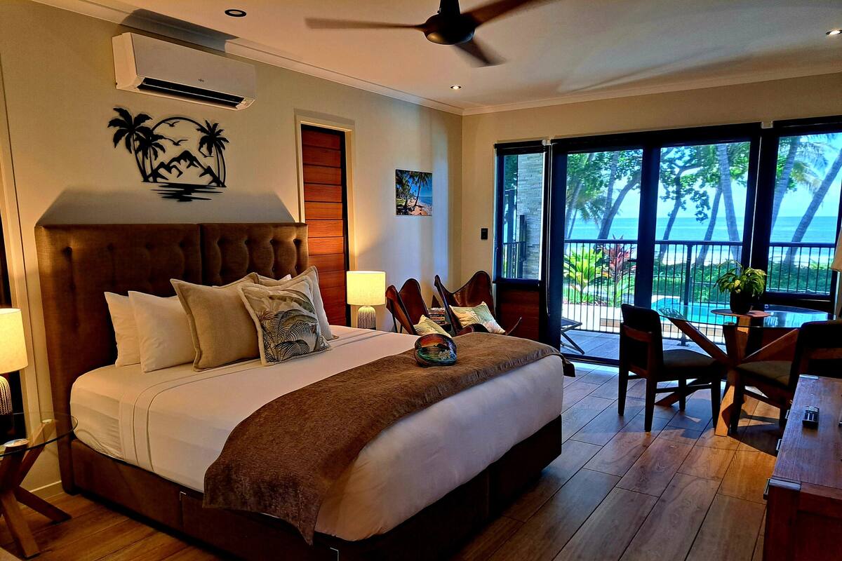 A bedroom with a large bed, a ceiling fan, a nightstand, a lamp, a window with a view of the outdoors, and a small table with chairs.