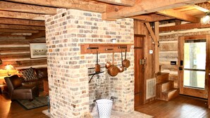 A rustic log cabin interior with a brick fireplace, wooden beams, a leather sofa, and a dining table.