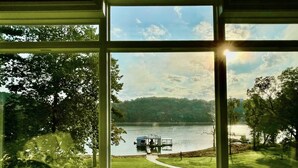 Une vue à travers une fenêtre sur un lac, avec un quai et un bateau, le tout entouré d’arbres et d’une zone herbeuse.