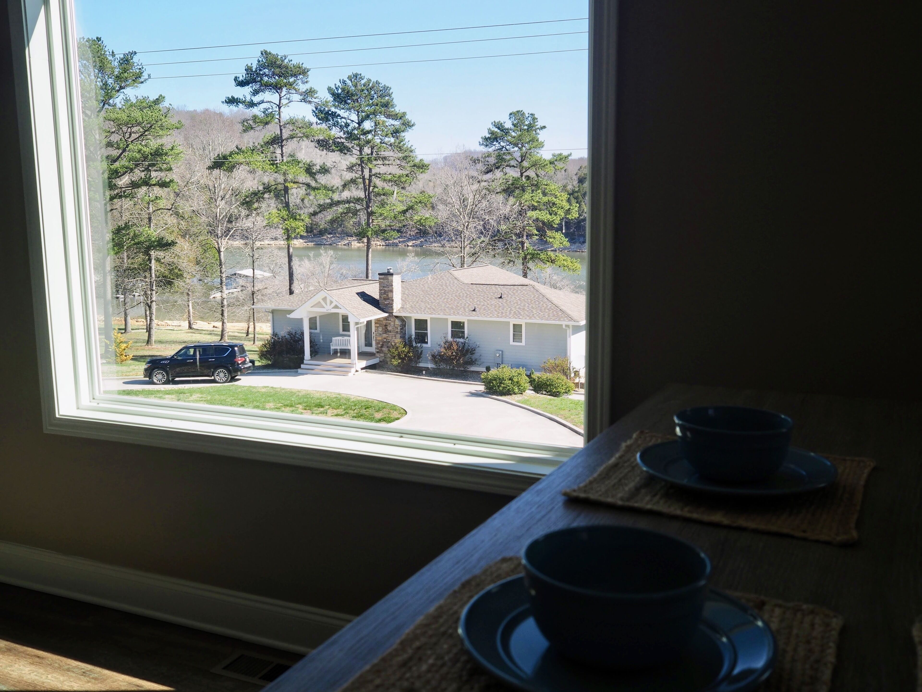 A view from a window overlooking a house with a driveway, a parked car, and a lake in the background.