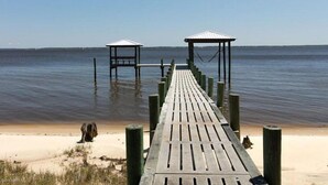 Eine Holzstege führt zu zwei kleinen Pavillons an einem sandigen Strand, im Hintergrund liegt ruhiges Wasser.