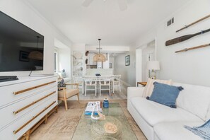 Ein modernes Wohnzimmer mit einem weißen Sofa, einem Flachbildfernseher, einer Essecke mit weißem Tisch und einer Küche im Hintergrund.